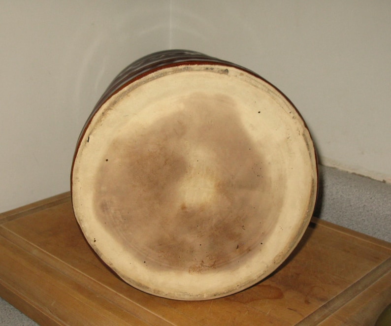 May include: A brown ceramic jug with a wide, circular base and a cylindrical body. The jug has a light brown interior and a darker brown exterior with horizontal ridges. The jug is sitting on a wooden surface.