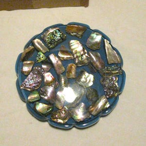 May include: A round, blue decorative object adorned with iridescent abalone shells. The shells are various shapes and sizes, reflecting light with colors of green, silver, and gold. The object has a scalloped edge.
