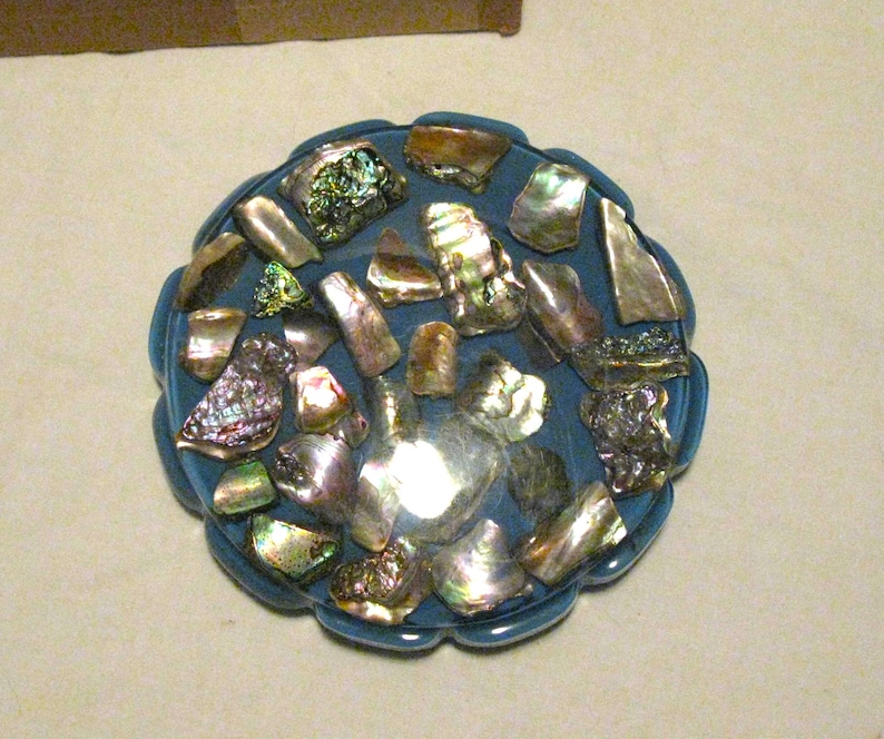 May include: A circular, blue decorative item featuring iridescent abalone shells. The shells, in diverse shapes and sizes, reflect light with green, silver, and gold hues. The item has a scalloped edge, adding to its design.