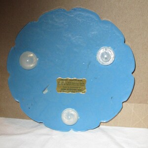 May include: A blue trivet with a scalloped edge and three clear, rounded feet. A small, rectangular label is affixed to the trivet. The trivet is designed to protect surfaces from hot dishes.