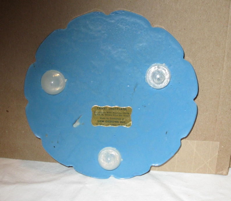 May include: A blue trivet with a scalloped edge and three clear, rounded feet. A small, rectangular label is affixed to the trivet. The trivet is designed to protect surfaces from hot dishes.