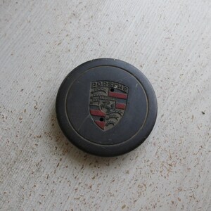 Puede incluir: Un tapacubos Porsche gris oscuro con el emblema de Porsche en el centro. El emblema presenta la palabra "PORSCHE" sobre un escudo con detalles rojos y negros. Se ven dos peque&ntilde;os agujeros debajo.