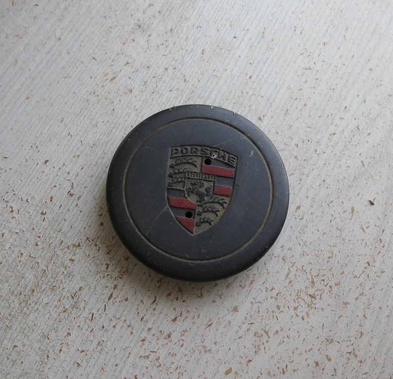 Puede incluir: Un tapacubos Porsche gris oscuro con el emblema de Porsche en el centro. El emblema presenta la palabra "PORSCHE" sobre un escudo con detalles rojos y negros. Se ven dos peque&ntilde;os agujeros debajo.