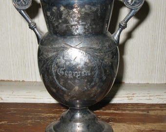 May include: A tarnished silver-toned trophy cup featuring two ornate handles with circular accents. The body of the cup displays an engraved laurel wreath design surrounding the word Georgia in a script font, set on a circular pedestal base.