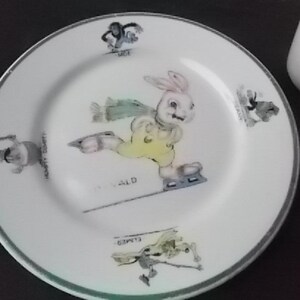 May include: White ceramic plate with a green rim featuring a cartoon rabbit ice skating. The rabbit wears a yellow outfit and skates on a line. The plate has illustrations of other characters and text around the rim.