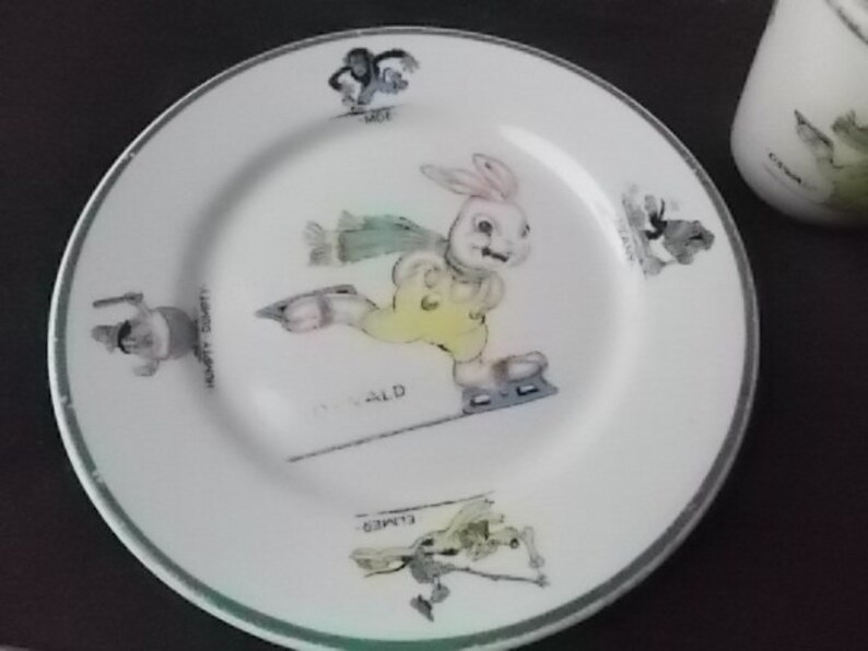 May include: White ceramic plate with a green rim featuring a cartoon rabbit ice skating. The rabbit wears a yellow outfit and skates on a line. The plate has illustrations of other characters and text around the rim.
