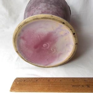 May include: A vintage ceramic vase with a speckled purple exterior and a pink and cream interior. The base has a circular design with a light beige rim. A wooden ruler is at the bottom.