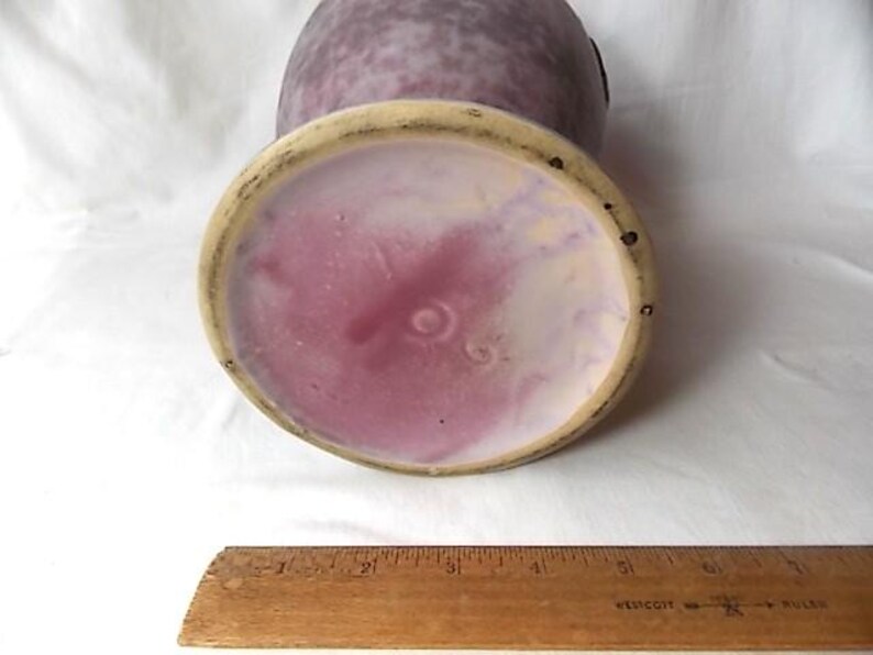 May include: A vintage ceramic vase with a speckled purple exterior and a pink and cream interior. The base has a circular design with a light beige rim. A wooden ruler is at the bottom.