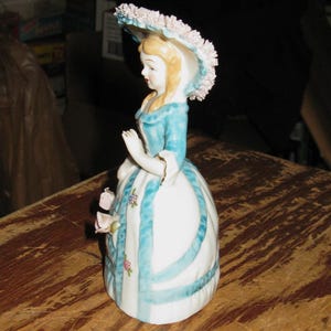 May include: A porcelain figurine of a person wearing a wide-brimmed hat with pink floral details, a blue bodice, and a white skirt featuring blue vertical stripes and small floral accents. The figure has blonde hair and is posed with one arm raised.