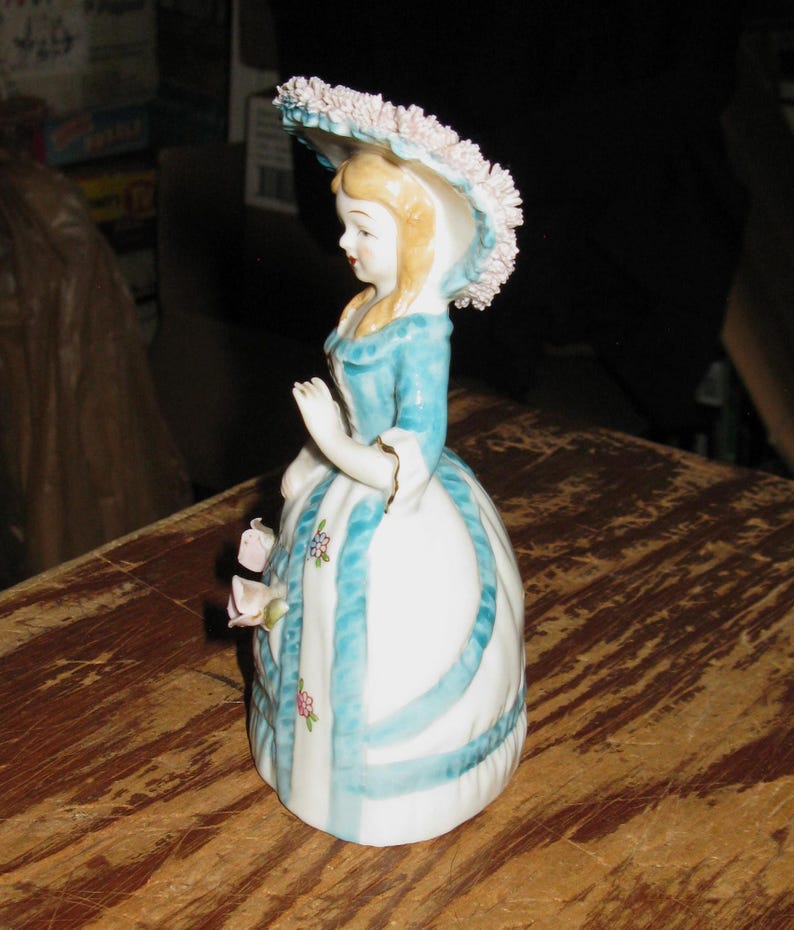 May include: A porcelain figurine of a person wearing a wide-brimmed hat with pink floral details, a blue bodice, and a white skirt featuring blue vertical stripes and small floral accents. The figure has blonde hair and is posed with one arm raised.