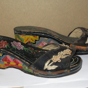 Peut inclure: Une paire de sandales &agrave; plateforme noires avec des bouts ouverts et des d&eacute;tails floraux brod&eacute;s. Les talons compens&eacute;s sont d&eacute;cor&eacute;s de sc&egrave;nes peintes &agrave; la main repr&eacute;sentant des b&acirc;timents et du feuillage en jaune, rouge et bleu.