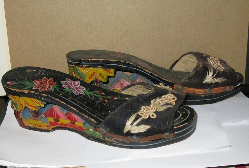 Peut inclure: Une paire de sandales &agrave; plateforme noires avec des bouts ouverts et des d&eacute;tails floraux brod&eacute;s. Les talons compens&eacute;s sont d&eacute;cor&eacute;s de sc&egrave;nes peintes &agrave; la main repr&eacute;sentant des b&acirc;timents et du feuillage en jaune, rouge et bleu.