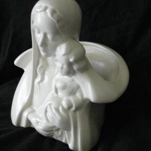 May include: A white ceramic sculpture of the Virgin Mary holding a child. The sculpture has a smooth, glossy finish and is set against a dark background. The figures are draped in flowing robes.