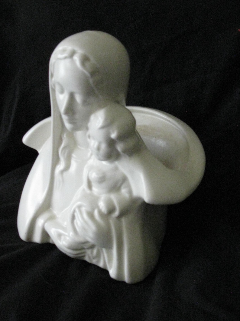 May include: A white ceramic sculpture of the Virgin Mary holding a child. The sculpture has a smooth, glossy finish and is set against a dark background. The figures are draped in flowing robes.