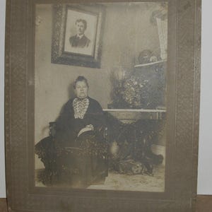 May include: A vintage black and white photograph of a woman seated in a chair, wearing a dark dress with a lace collar. A framed portrait hangs on the wall behind her. The photograph is in a decorative frame.