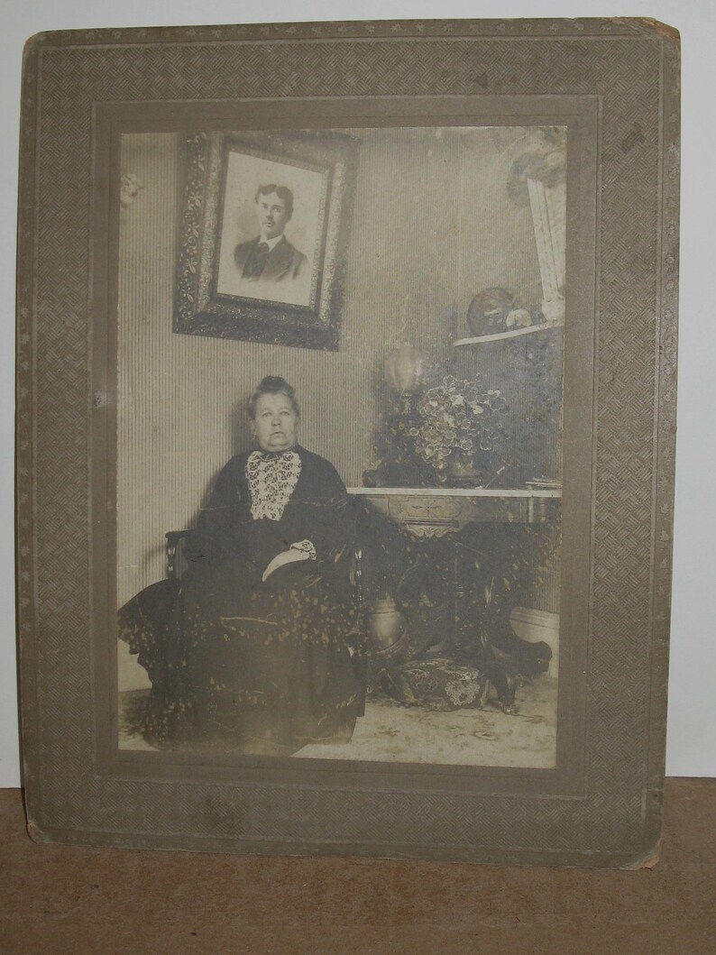 May include: A vintage black and white photograph of a woman seated in a chair, wearing a dark dress with a lace collar. A framed portrait hangs on the wall behind her. The photograph is in a decorative frame.