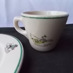 May include: Cream-colored teacup and saucer set with a green trim. The teacup features a cartoon illustration of a person fishing, with the name "ELMER" below. The saucer has a similar illustration and green trim, suggesting a vintage or collectible theme.