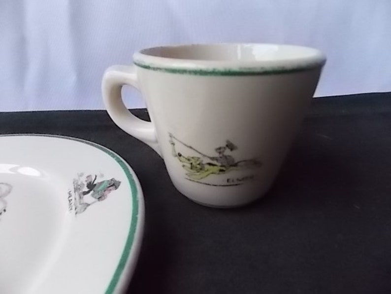 May include: Cream-colored teacup and saucer set with a green trim. The teacup features a cartoon illustration of a person fishing, with the name "ELMER" below. The saucer has a similar illustration and green trim, suggesting a vintage or collectible theme.