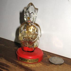 K&ouml;nnte beinhalten: Eine klare Glasfigur einer sitzenden elfenartigen Figur mit rotem Sockel. Die Figur hat ein bemaltes Gesicht mit roter Nase und Mund. Der Sockel tr&auml;gt die Aufschrift "DEVON VIOLETS".