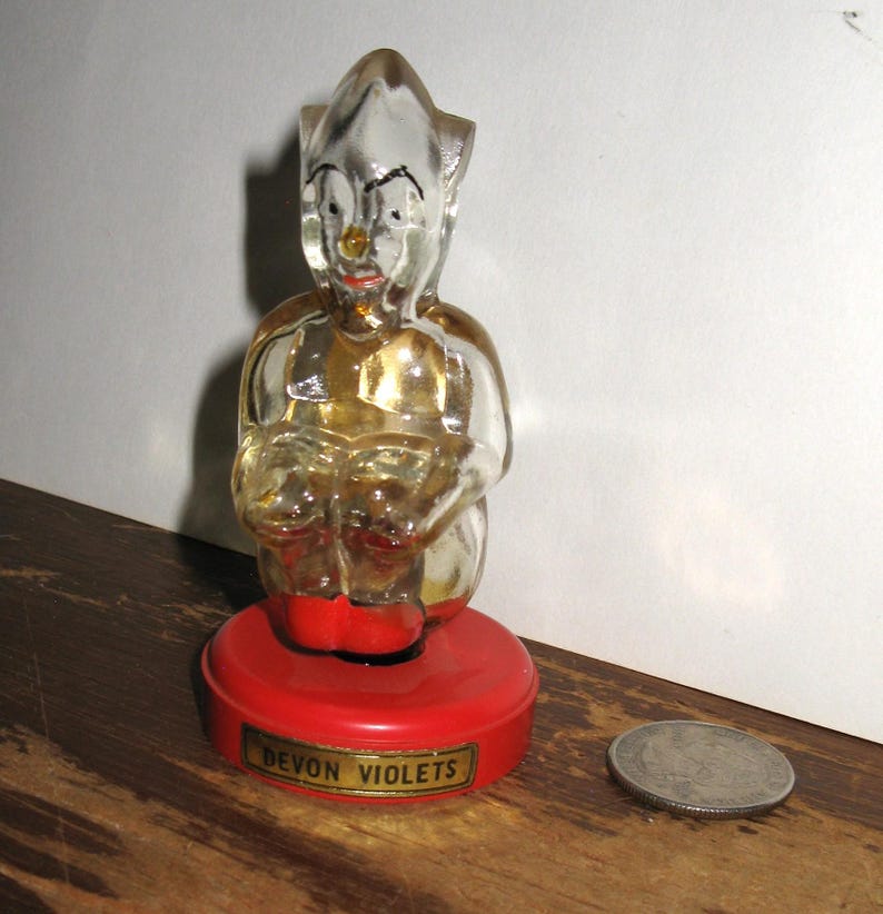 K&ouml;nnte beinhalten: Eine klare Glasfigur einer sitzenden elfenartigen Figur mit rotem Sockel. Die Figur hat ein bemaltes Gesicht mit roter Nase und Mund. Der Sockel tr&auml;gt die Aufschrift "DEVON VIOLETS".