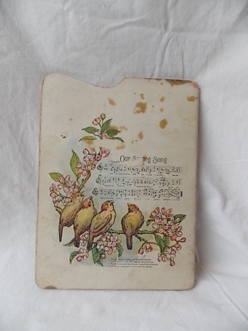 Puede incluir: Una tarjeta rectangular de color blanco roto con esquinas superiores redondeadas que presenta una ilustraci&oacute;n impresa de cuatro p&aacute;jaros posados en una rama florida. El dise&ntilde;o incluye notaci&oacute;n musical y el texto Our S-ing Song, con manchas.