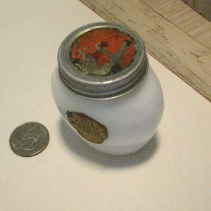 May include: A small, white, oval-shaped jar with a silver lid and a decorative label. The lid features a colorful image, and a gold-toned label is on the side. A quarter is next to the jar for size comparison.