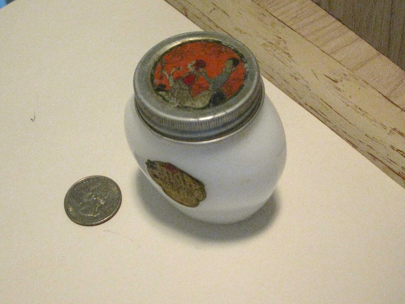 May include: A small, white, oval-shaped jar with a silver lid and a decorative label. The lid features a colorful image, and a gold-toned label is on the side. A quarter is next to the jar for size comparison.