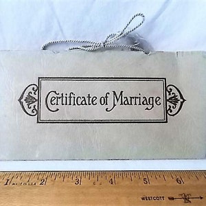 May include: A cream-colored Certificate of Marriage with black text and decorative flourishes. The certificate is rectangular with a string attached at the top. A wooden ruler with inch measurements is at the bottom.