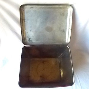 May include: A rectangular metal storage tin with an attached hinged lid. The interior of the container and the underside of the lid feature a weathered, metallic finish with visible signs of oxidation and discoloration throughout the surface.