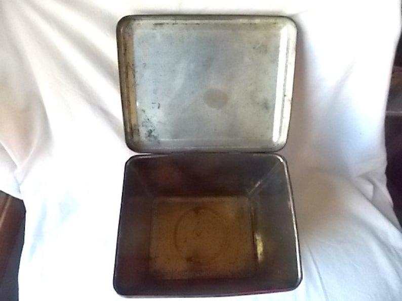 May include: A rectangular metal storage tin with an attached hinged lid. The interior of the container and the underside of the lid feature a weathered, metallic finish with visible signs of oxidation and discoloration throughout the surface.