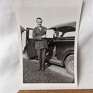 May include: Black and white photograph of a man in a military uniform standing next to a vintage car. The man is holding a document and smiling. The car door is open, and the background shows a grassy area and a building.