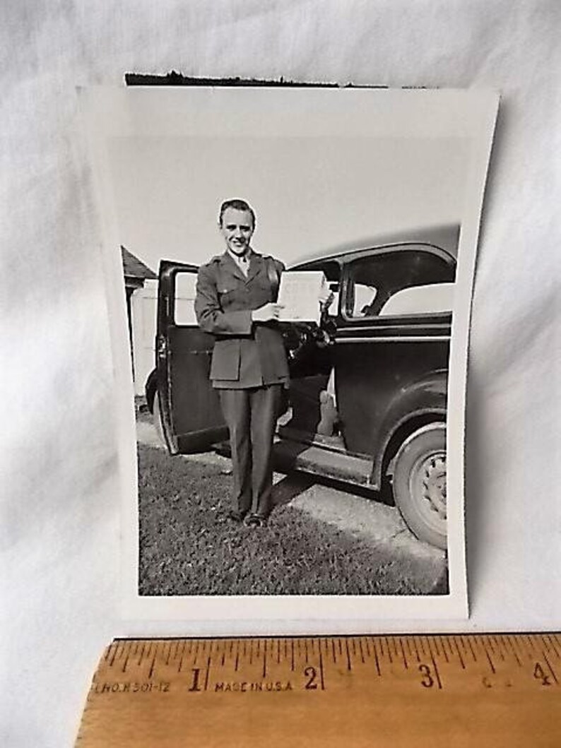 May include: Black and white photograph of a man in a military uniform standing next to a vintage car. The man is holding a document and smiling. The car door is open, and the background shows a grassy area and a building.