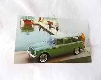 May include: A vintage postcard featuring a light green station wagon parked on a concrete dock next to a body of water. A person in a yellow shirt stands by the rear of the vehicle, while other figures walk along the pier toward a boat.
