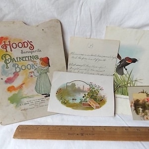Puede incluir: Una colecci&oacute;n de ef&iacute;meros de papel vintage que incluye un libro para colorear Hood's Sarsaparilla, una nota manuscrita, una ilustraci&oacute;n de un p&aacute;jaro y una tarjeta floral, dispuestos sobre una regla de madera de 30 cm en una superficie blanca.
