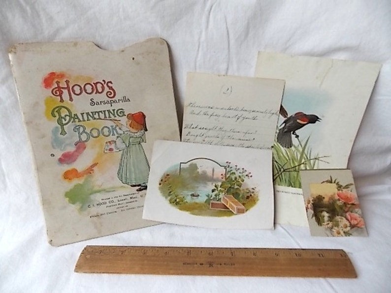 Puede incluir: Una colecci&oacute;n de ef&iacute;meros de papel vintage que incluye un libro para colorear Hood's Sarsaparilla, una nota manuscrita, una ilustraci&oacute;n de un p&aacute;jaro y una tarjeta floral, dispuestos sobre una regla de madera de 30 cm en una superficie blanca.