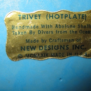 May include: A blue trivet with a gold label. The label reads "TRIVET (HOTPLATE) Handmade With Abalone Shells Taken By Divers from the Ocean Made by Craftsmen of NEW DESIGNS INC. MCMLXIX MADE IN U.S.A."