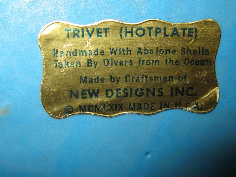 May include: A blue trivet with a gold label. The label reads "TRIVET (HOTPLATE) Handmade With Abalone Shells Taken By Divers from the Ocean Made by Craftsmen of NEW DESIGNS INC. MCMLXIX MADE IN U.S.A."