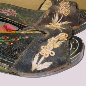 Peut inclure: Une paire de sandales &agrave; plateforme en bois noir avec des brides en tissu sombre orn&eacute;es de broderies florales claires. Les semelles sont peintes de motifs floraux color&eacute;s et de formes rouges rappelant des montagnes, avec des points jaunes.