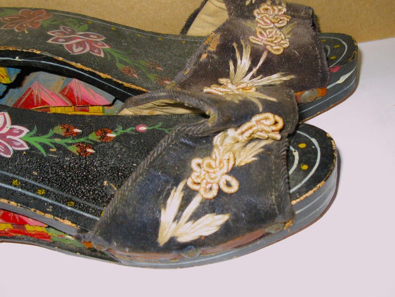 Peut inclure: Une paire de sandales &agrave; plateforme en bois noir avec des brides en tissu sombre orn&eacute;es de broderies florales claires. Les semelles sont peintes de motifs floraux color&eacute;s et de formes rouges rappelant des montagnes, avec des points jaunes.