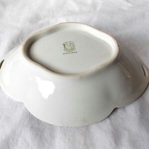 May include: A white porcelain dish with a scalloped edge and two small handles. The dish has a slightly raised rim and a smooth, glossy surface. The base of the dish has a green and gold emblem and the words "Made in Japan".