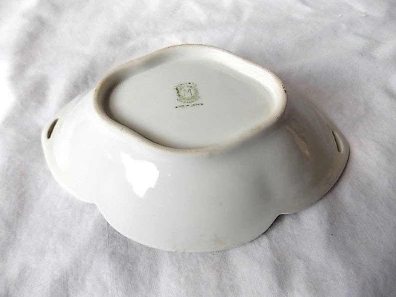 May include: A white porcelain dish with a scalloped edge and two small handles. The dish has a slightly raised rim and a smooth, glossy surface. The base of the dish has a green and gold emblem and the words "Made in Japan".