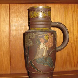 Peut inclure: Une grande cruche cylindrique en c&eacute;ramique brune orn&eacute;e d'un relief repr&eacute;sentant une personne assise tenant un pichet, avec une faux &agrave; proximit&eacute;. La cruche pr&eacute;sente une base brun fonc&eacute;, une anse incurv&eacute;e et une bande d&eacute;corative verte et brune.