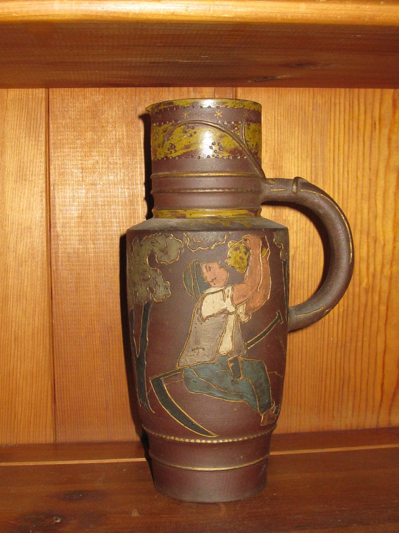 Peut inclure: Une grande cruche cylindrique en c&eacute;ramique brune orn&eacute;e d'un relief repr&eacute;sentant une personne assise tenant un pichet, avec une faux &agrave; proximit&eacute;. La cruche pr&eacute;sente une base brun fonc&eacute;, une anse incurv&eacute;e et une bande d&eacute;corative verte et brune.