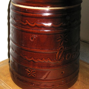 May include: A brown ceramic cookie jar with a lid. The jar has a cylindrical shape with horizontal bands and decorative wave and flower patterns. The word "Cookies" is written on the side.