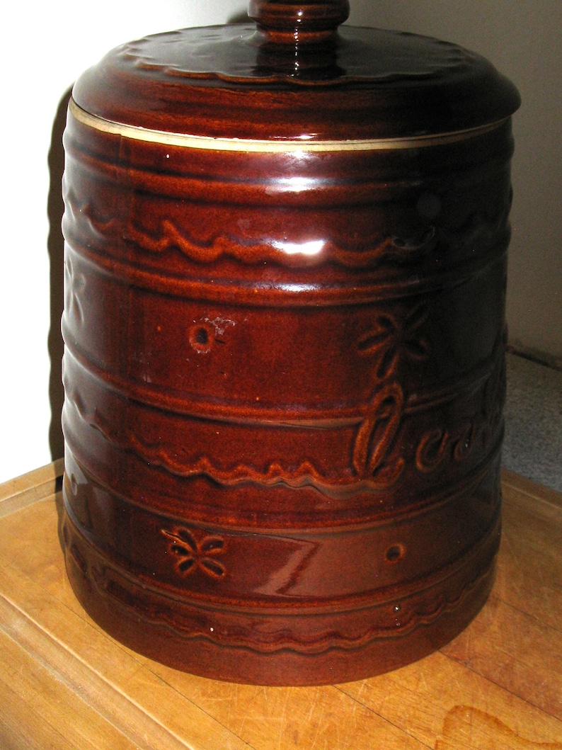 May include: A brown ceramic cookie jar with a lid. The jar has a cylindrical shape with horizontal bands and decorative wave and flower patterns. The word "Cookies" is written on the side.