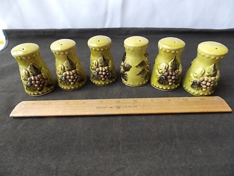 May include: Set of six ceramic salt and pepper shakers. The shakers are light green with a grape cluster design in brown and green. Each shaker has a small hole on top for dispensing and a rounded base.