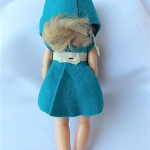 May include: A vintage doll wearing a teal felt dress and matching hood. The doll has blonde hair and a white belt. The doll's legs and feet are visible. The doll is a collectible item.
