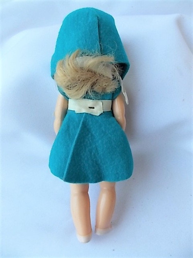 May include: A vintage doll wearing a teal felt dress and matching hood. The doll has blonde hair and a white belt. The doll's legs and feet are visible. The doll is a collectible item.