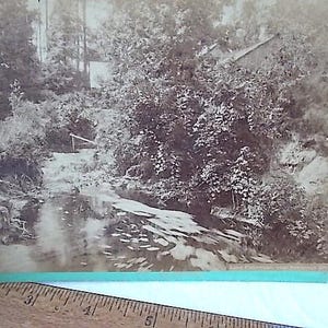 May include: Sepia-toned photograph of a serene outdoor scene. A person is fishing in a stream surrounded by lush trees and foliage. A small building is visible in the background, and a wooden structure spans the water.