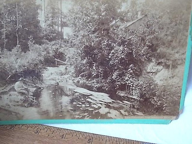 May include: Sepia-toned photograph of a serene outdoor scene. A person is fishing in a stream surrounded by lush trees and foliage. A small building is visible in the background, and a wooden structure spans the water.